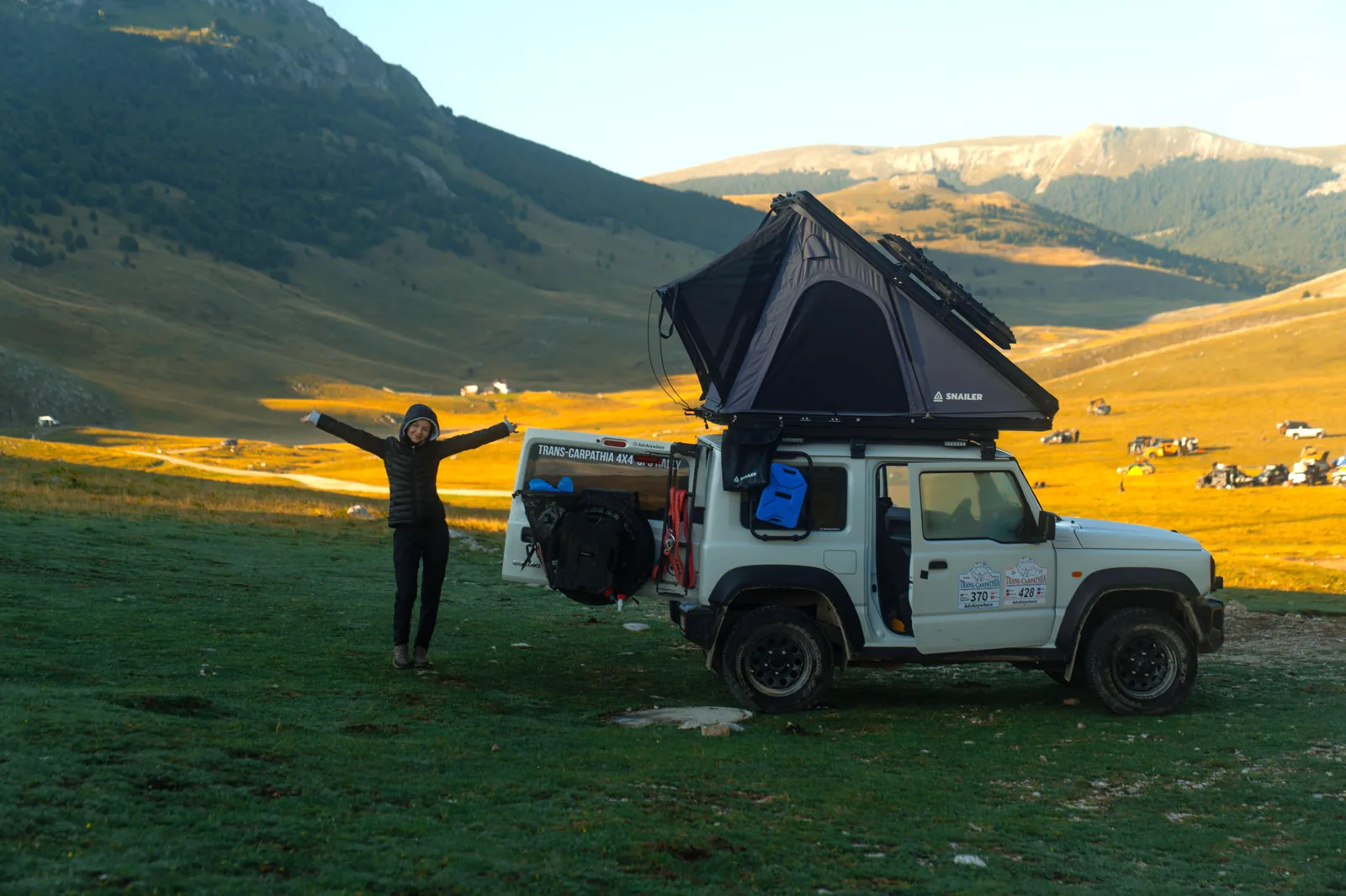 Viaggiatrice accanto a un fuoristrada con tenda da tetto SNAILER aperta in una valle di montagna
