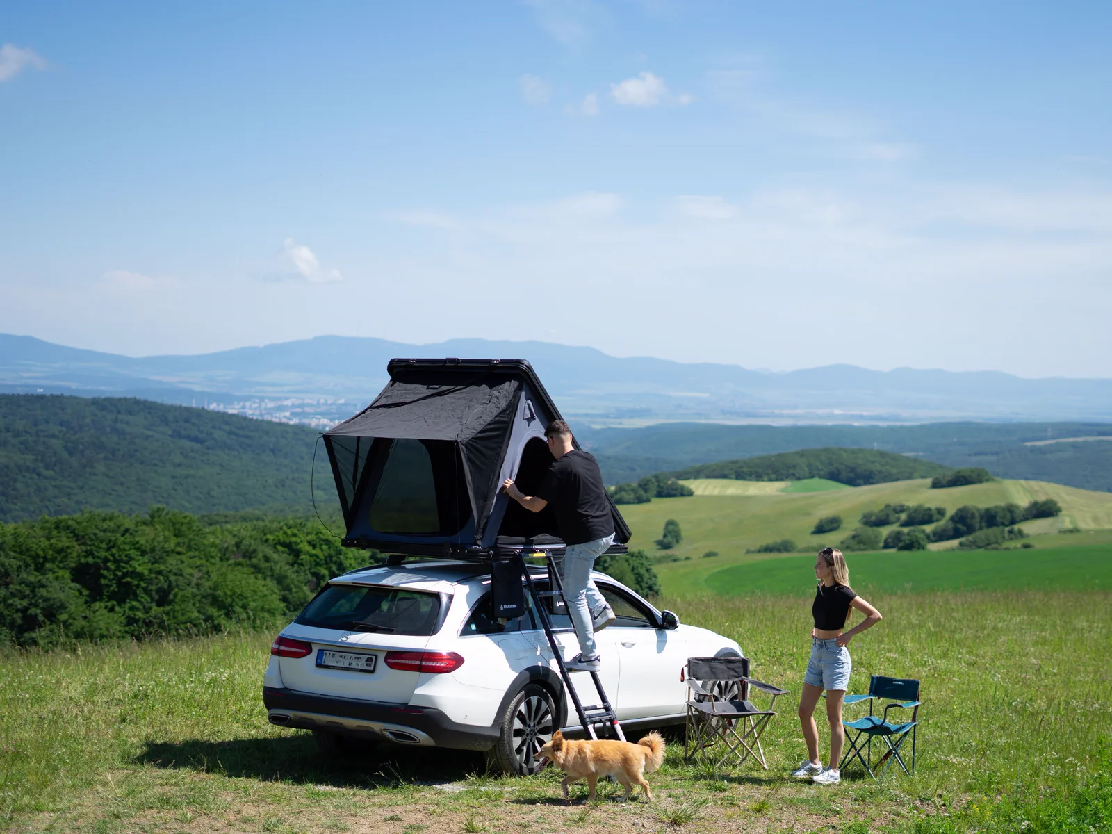 Una coppia apre una tenda da tetto SNAILER su un'auto in un prato di montagna